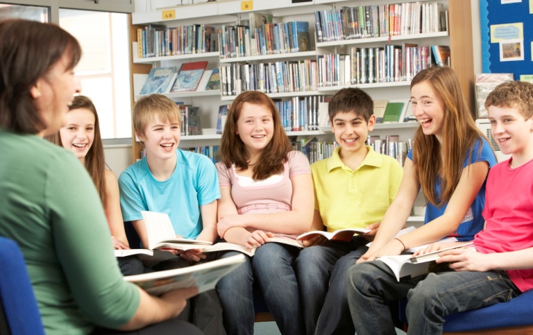 Lärare och sex barn i nedre tonåren med böcker i händerna pratar i bibliotek